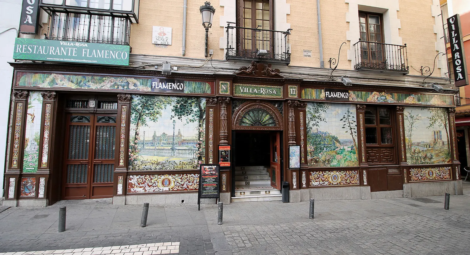The front of the former Villa Rosa tablao on Plaza de Santa Ana, a long wall of ceramic panels showing a plaza view, framed in dark carved wood, with a pale street and a corner door